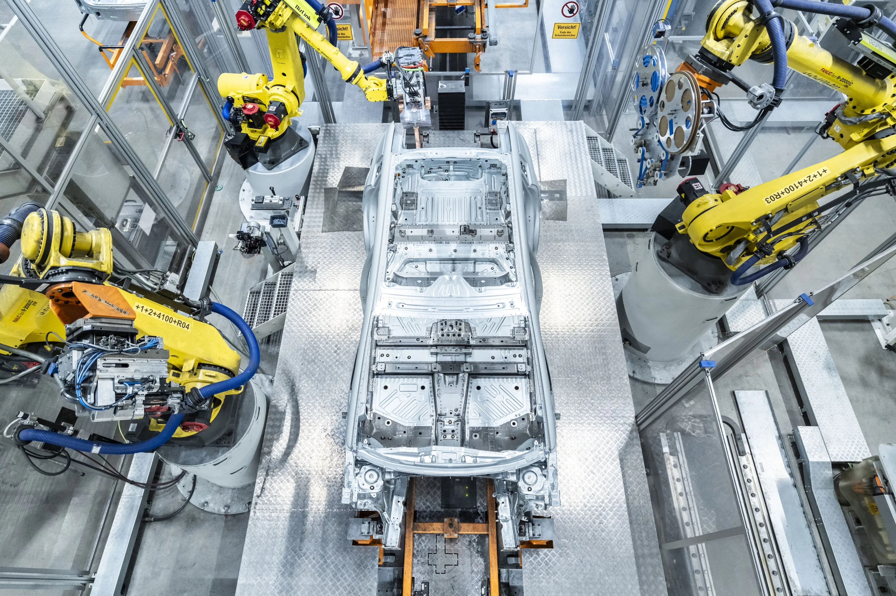 Eine Rohkarosserie zwischen gelben Industrierobotern in einer Fertigungszelle, von oben gesehen