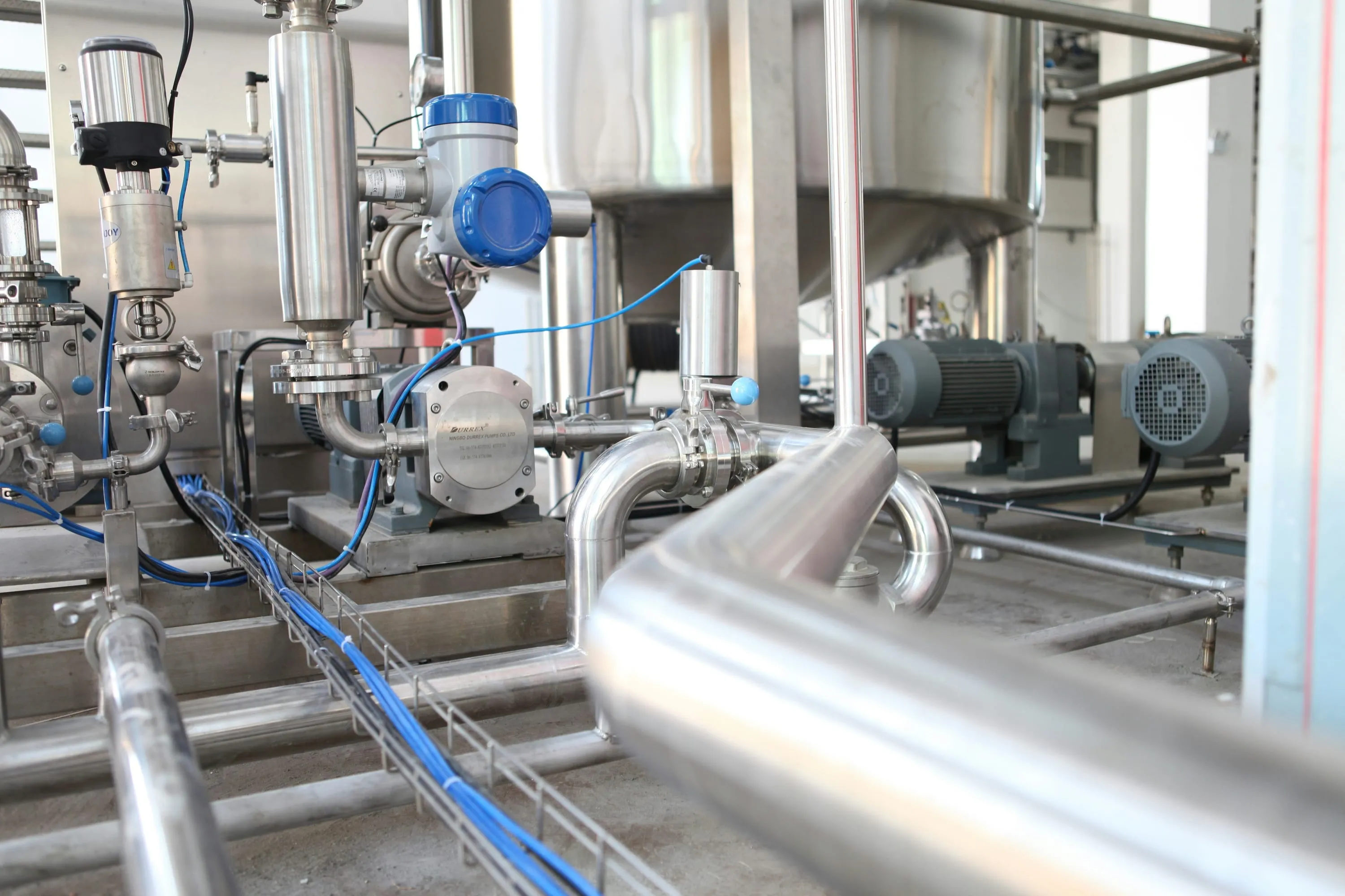 Edelstahlpumpen mit Motoren und Rohrleitungen vor Tanks in einer Prozessanlage