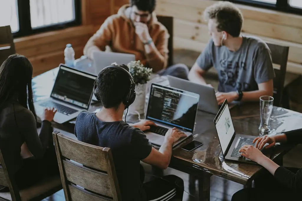 Fünf Personen arbeiten nebeneinander an einem gemeinsamen Tisch an ihren Laptops